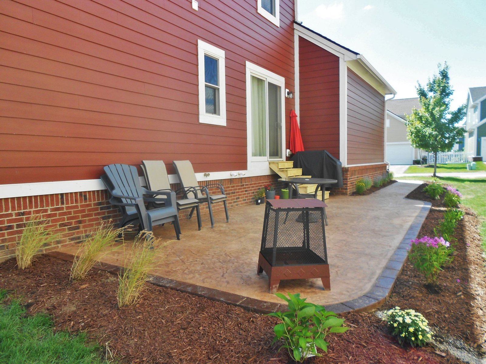 Stamped concrete patio with brick border behind a red-sided home, furnished with chairs and a fire pit