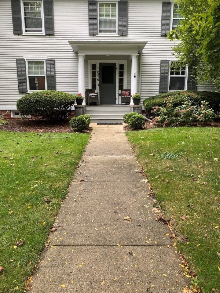 Front walkway to a white house with black shutters, green lawn, and a front porch with potted plants.