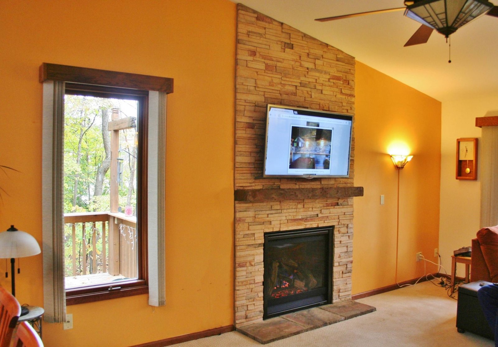 Living room with stone fireplace, mounted TV, orange walls, and a window by a wooden railing.