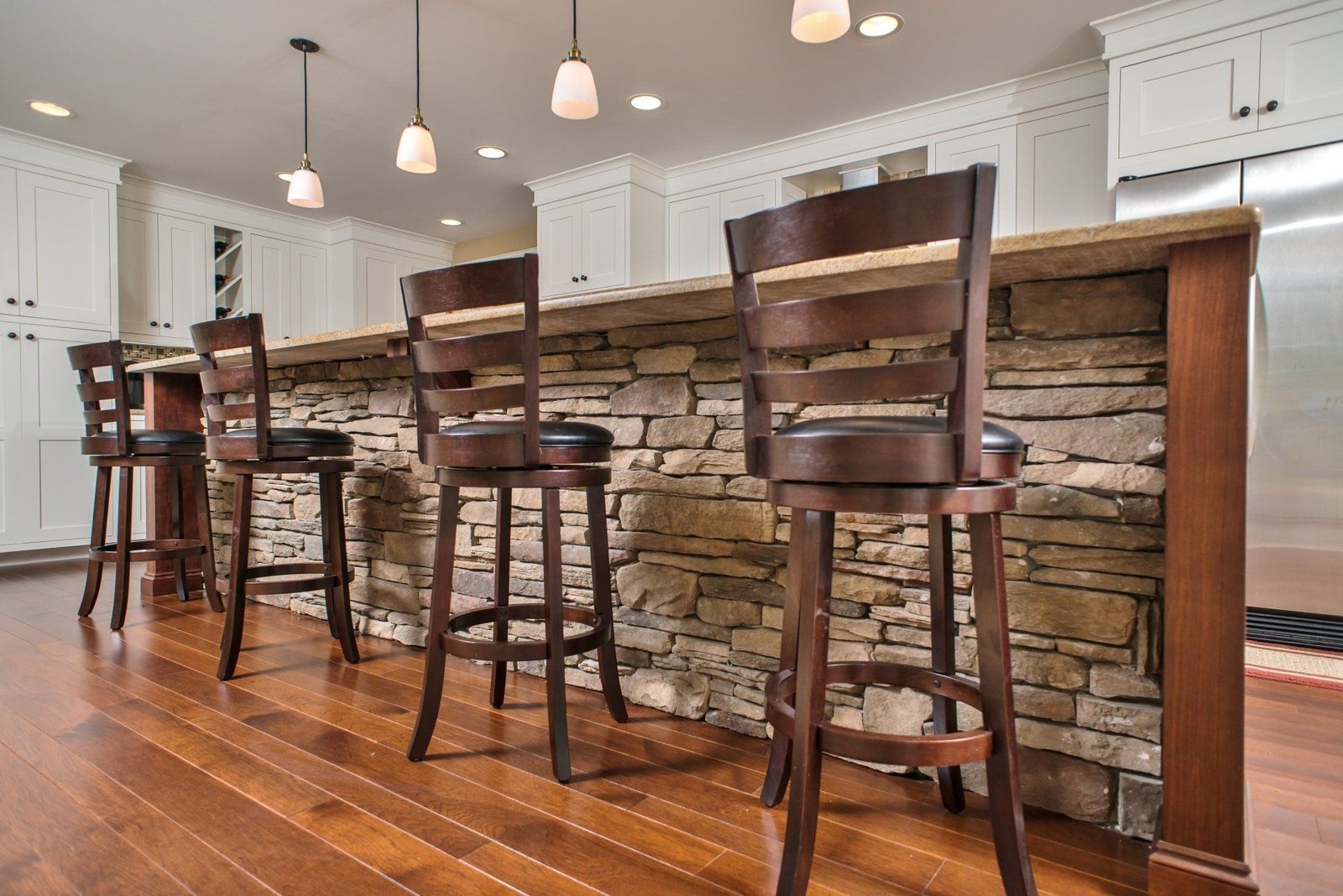 Kitchen island with stone front, three wooden bar stools, and warm hardwood floors
