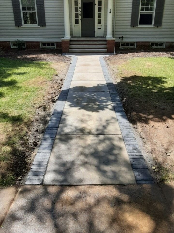 Concrete walkway leading to a house front steps, bordered by brick edges and patchy grass.