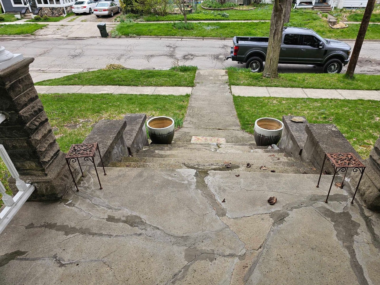 Front porch steps leading to a cracked concrete walkway, with potted plants and a street view beyond.