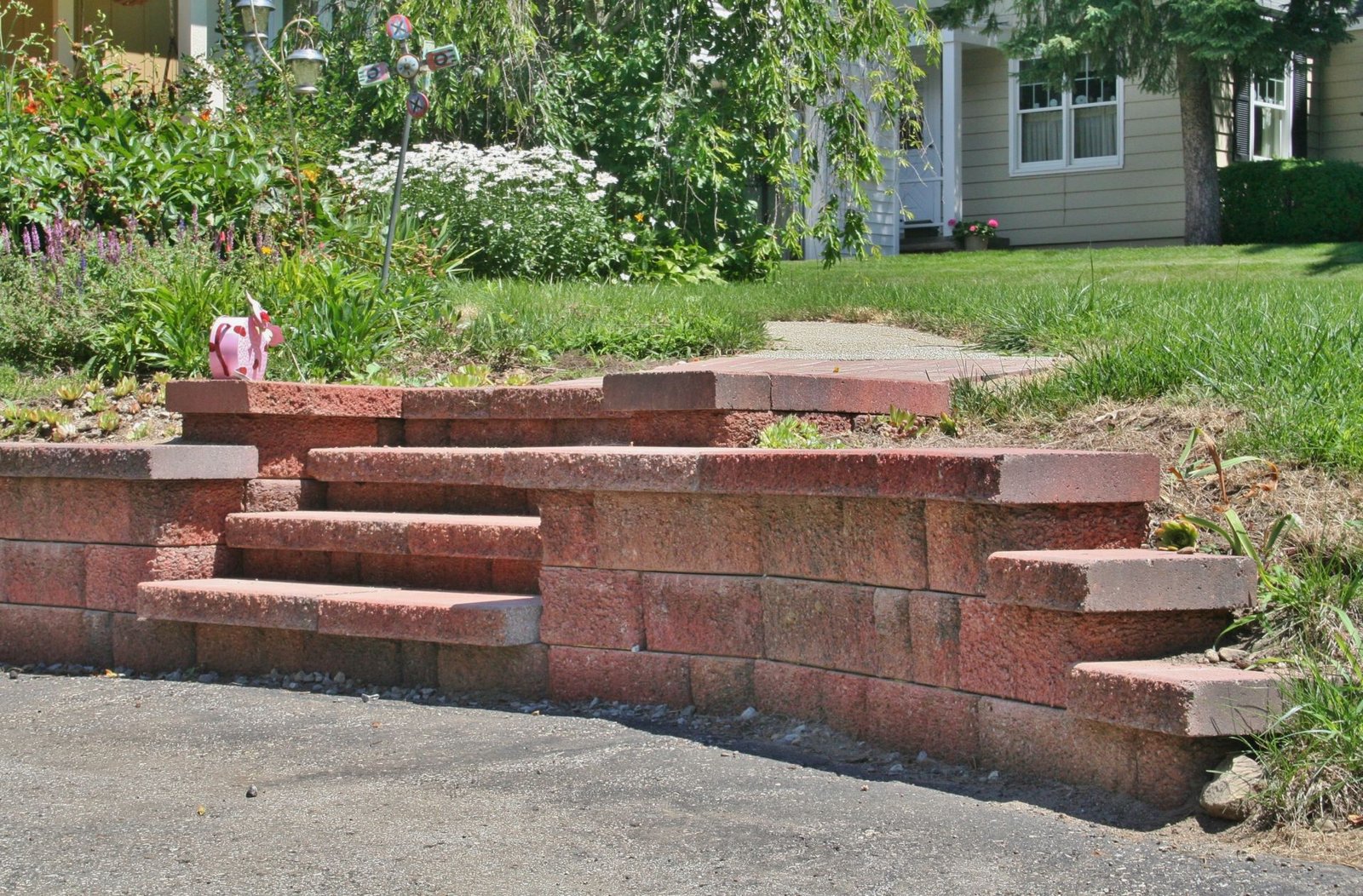 Brick retaining wall with steps beside a driveway and a grassy yard with flowers.