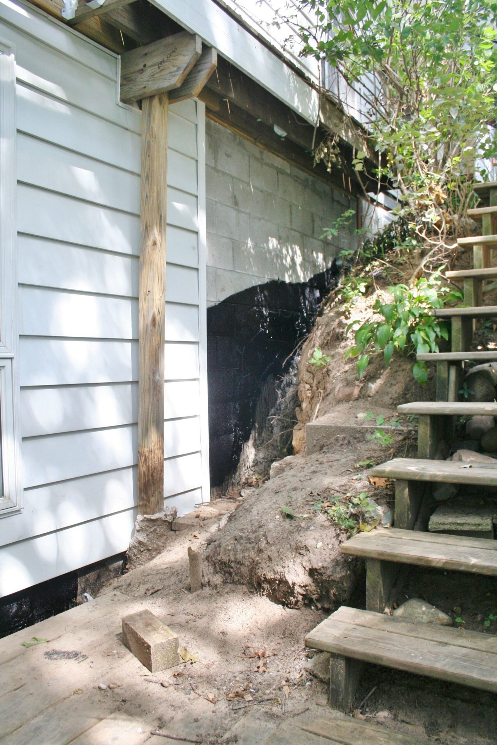 Exterior wall with exposed foundation and eroded soil beside wooden stairs and a retaining rock slope