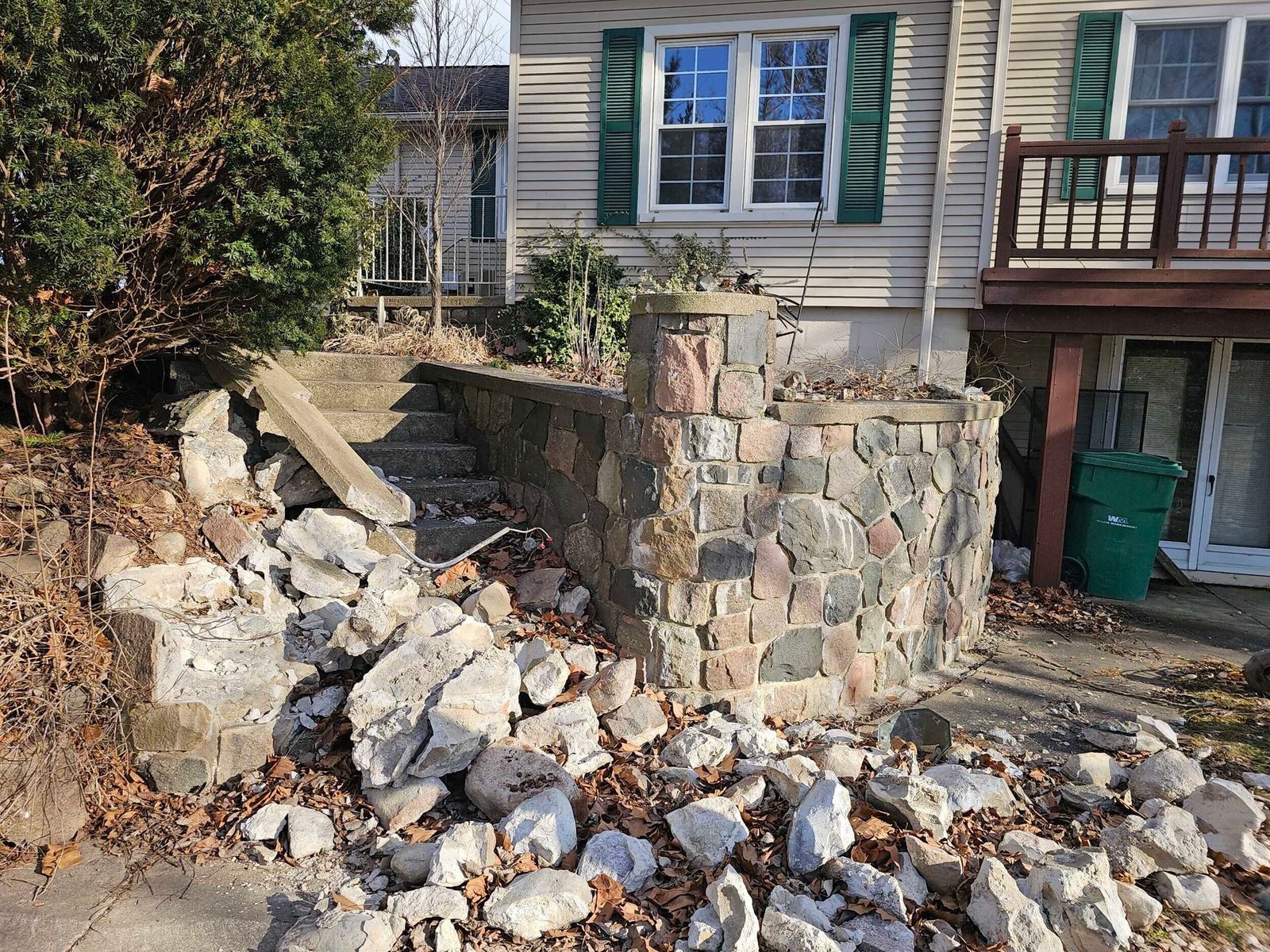 Collapsed stone retaining wall and rubble piled in front of a house with blue shutters.