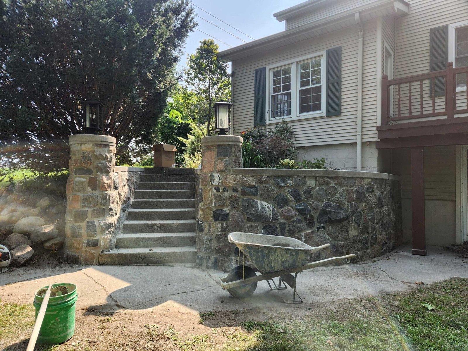 Front steps and stone patio beside a white house, with a wheelbarrow in the foreground.