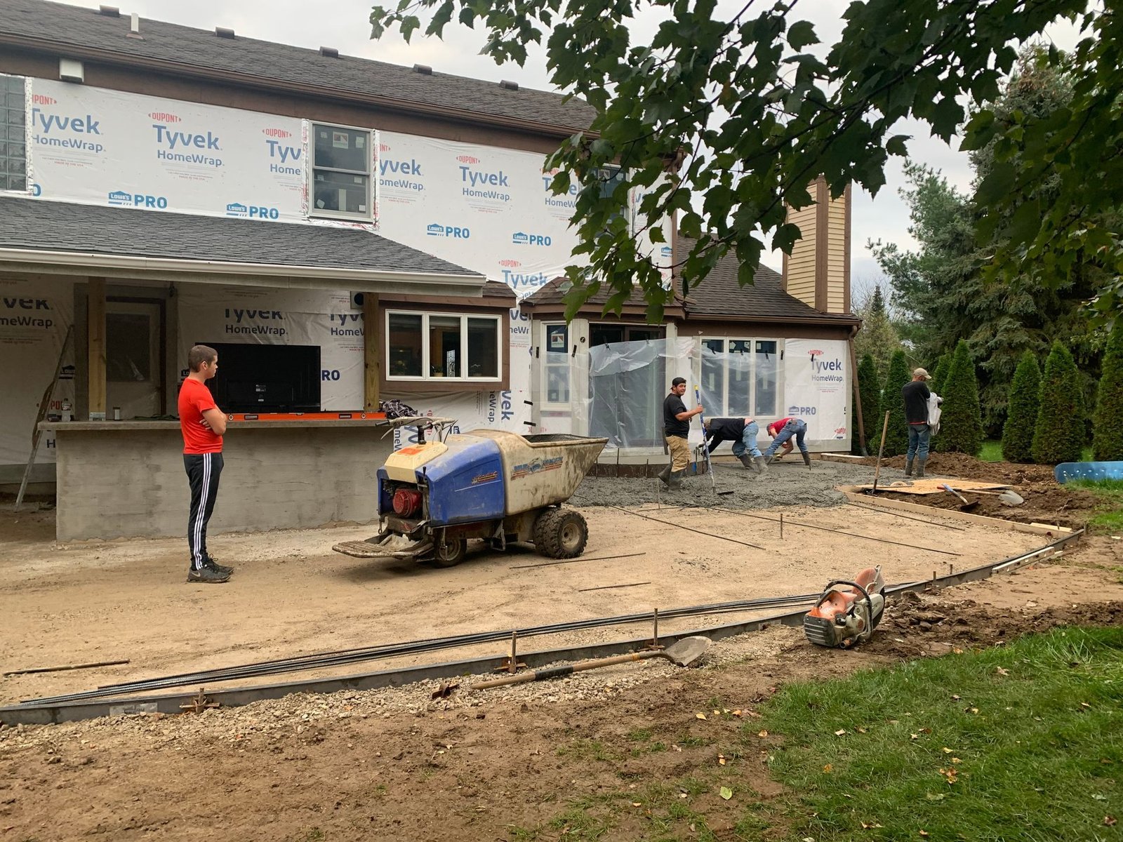 Construction site with workers, a small blue machine, and a house wrapped in building materials.