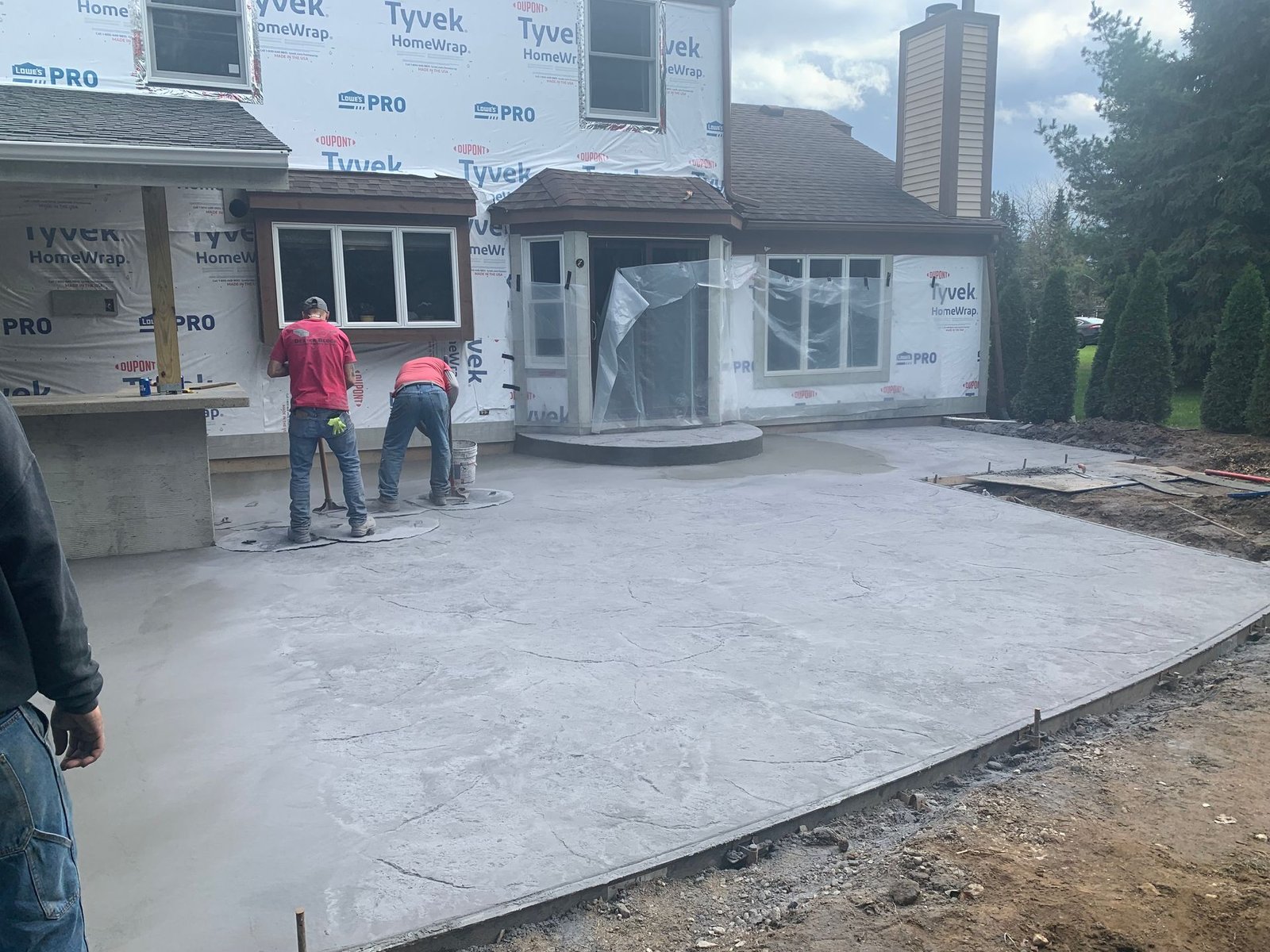 Two workers in red jackets stand on a newly poured concrete driveway beside a house under construction.