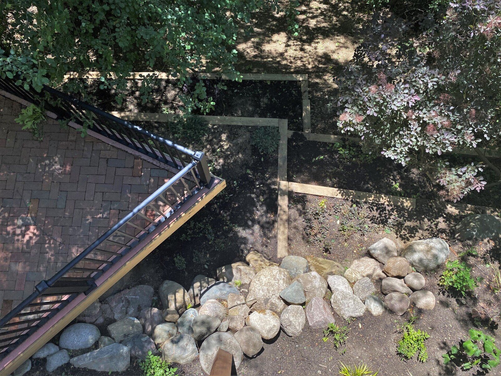 Stone-lined stream beneath a wooden bridge beside a brick path, shaded by trees.