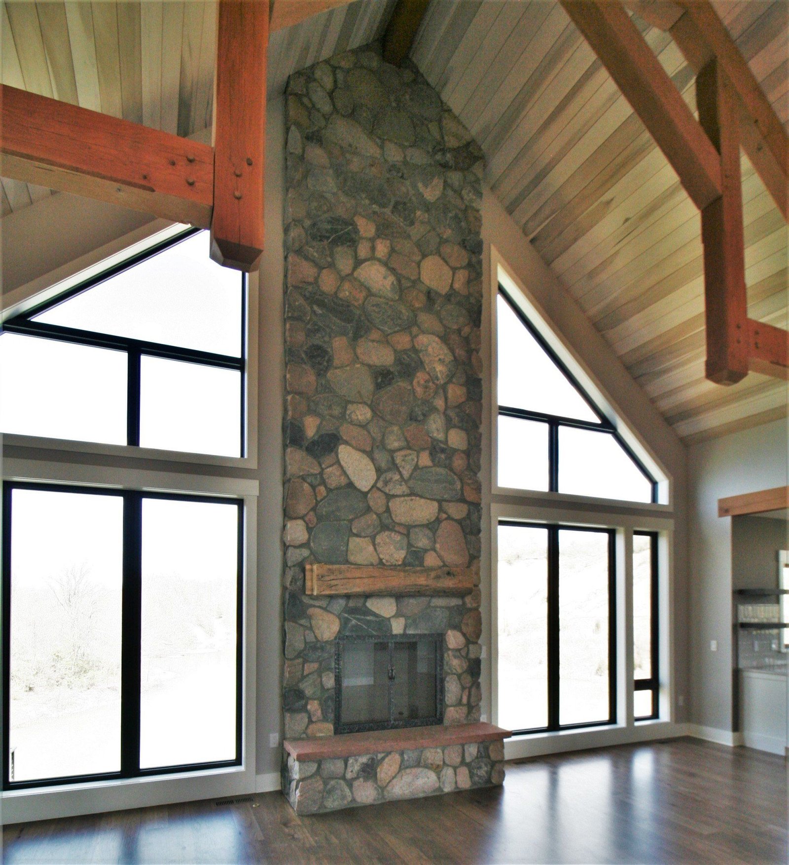 Stone fireplace in a vaulted room with large windows and exposed wooden beams.