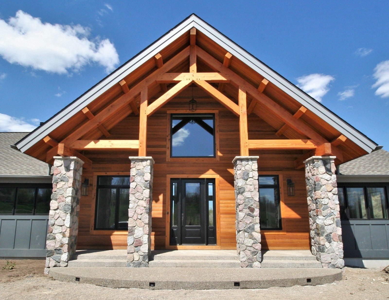 Front view of a modern rustic house with timber siding, stone pillars, and a peaked roof under a blue sky