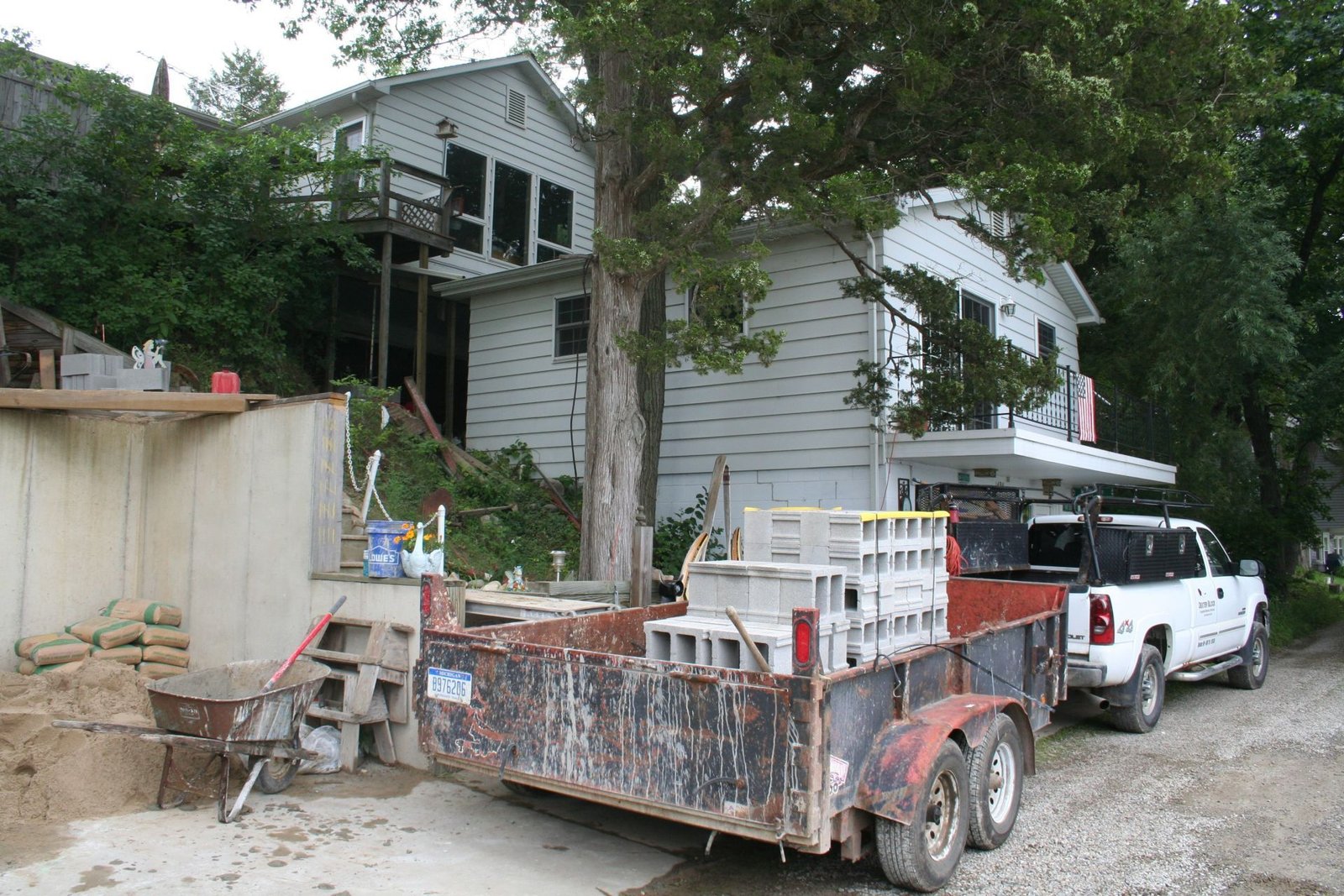 Old white house on a dirt lot with a rusty trailer and white pickup truck under trees