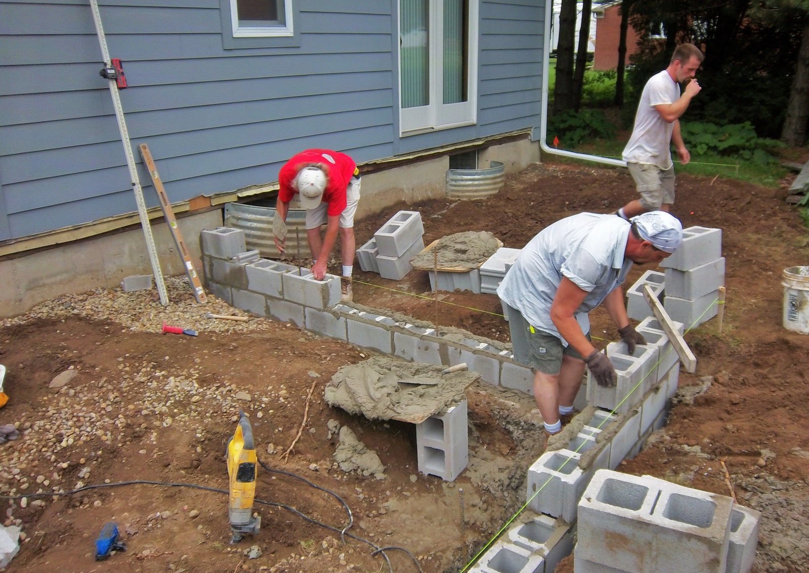 Workers laying cinder blocks to build a foundation beside a house.