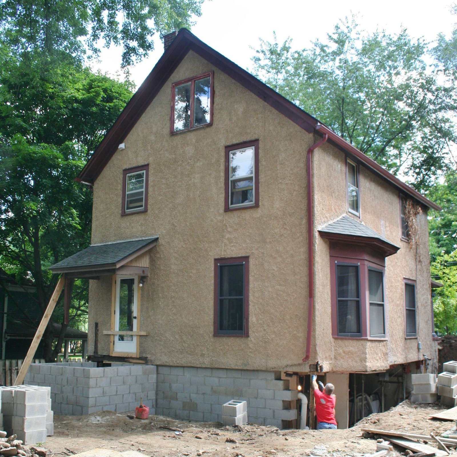 Partially renovated two-story tan house with exposed foundation and workers outside