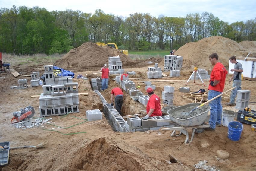Construction crew laying cinder blocks at a house foundation site, with wheelbarrow and dirt piles nearby