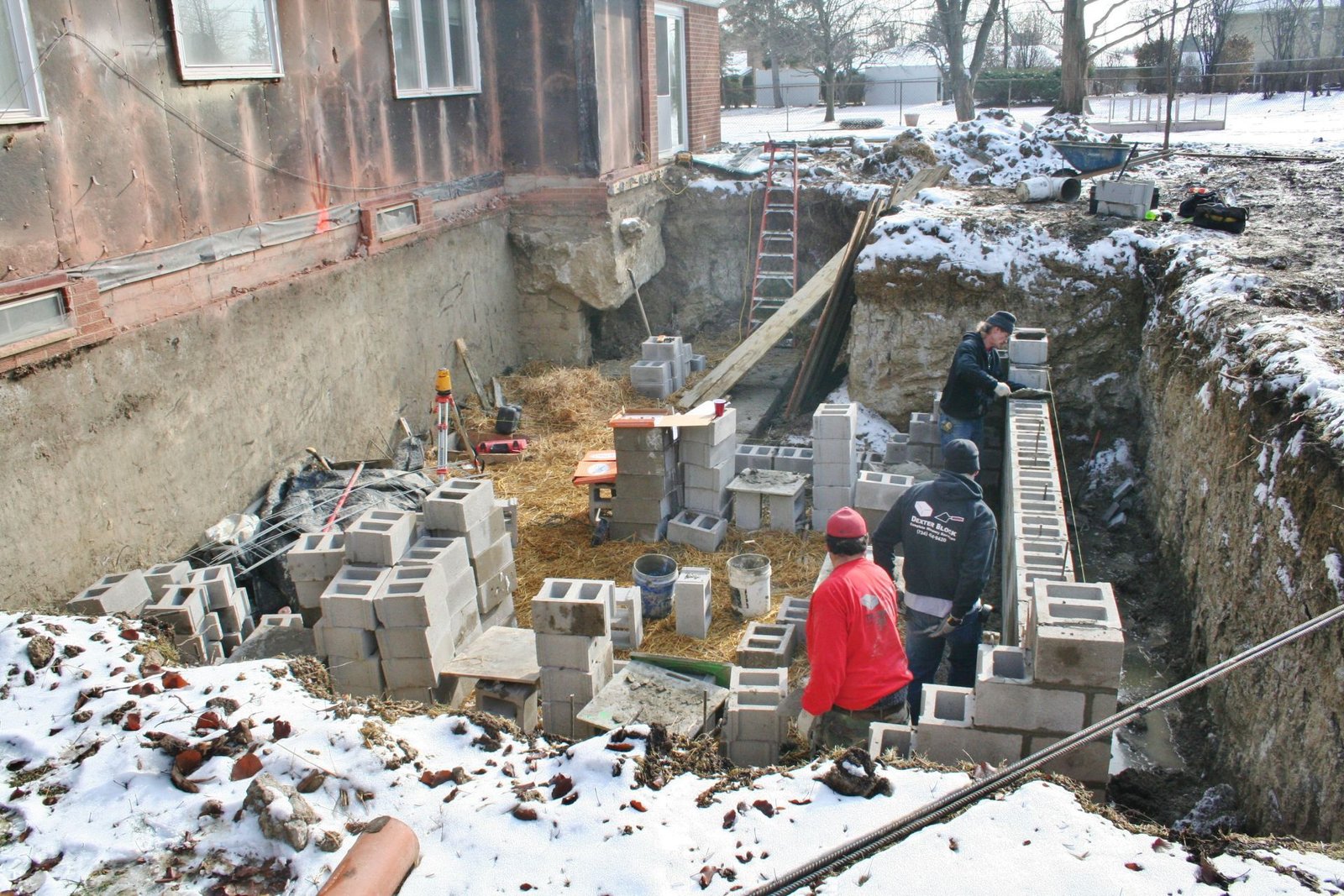 Snowy construction trench beside a building, with cinder blocks and two workers in red jackets.