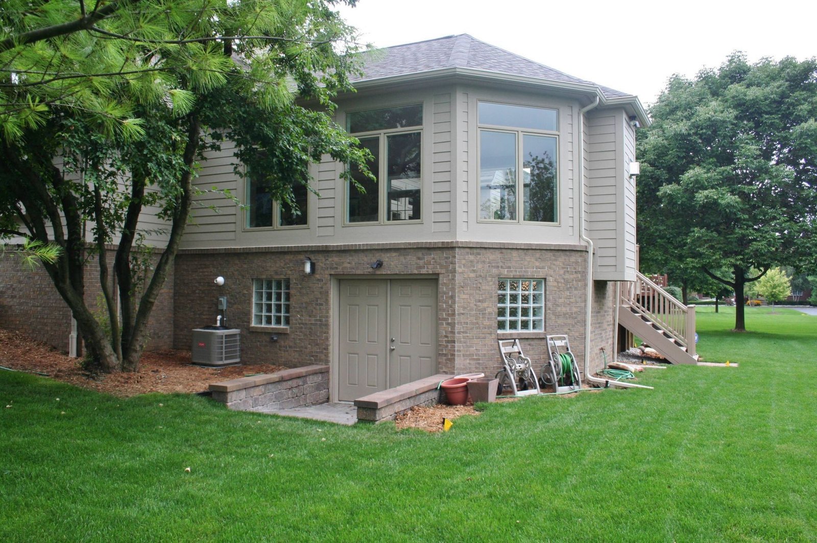 Two-story beige house with a large lawn, trees, and a garage beneath the main floor.