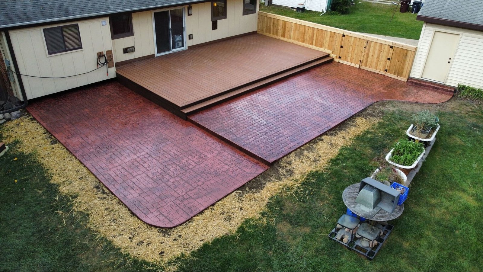 Backyard patio with maroon paving, a raised wooden deck, and a wheelbarrow on the grass.