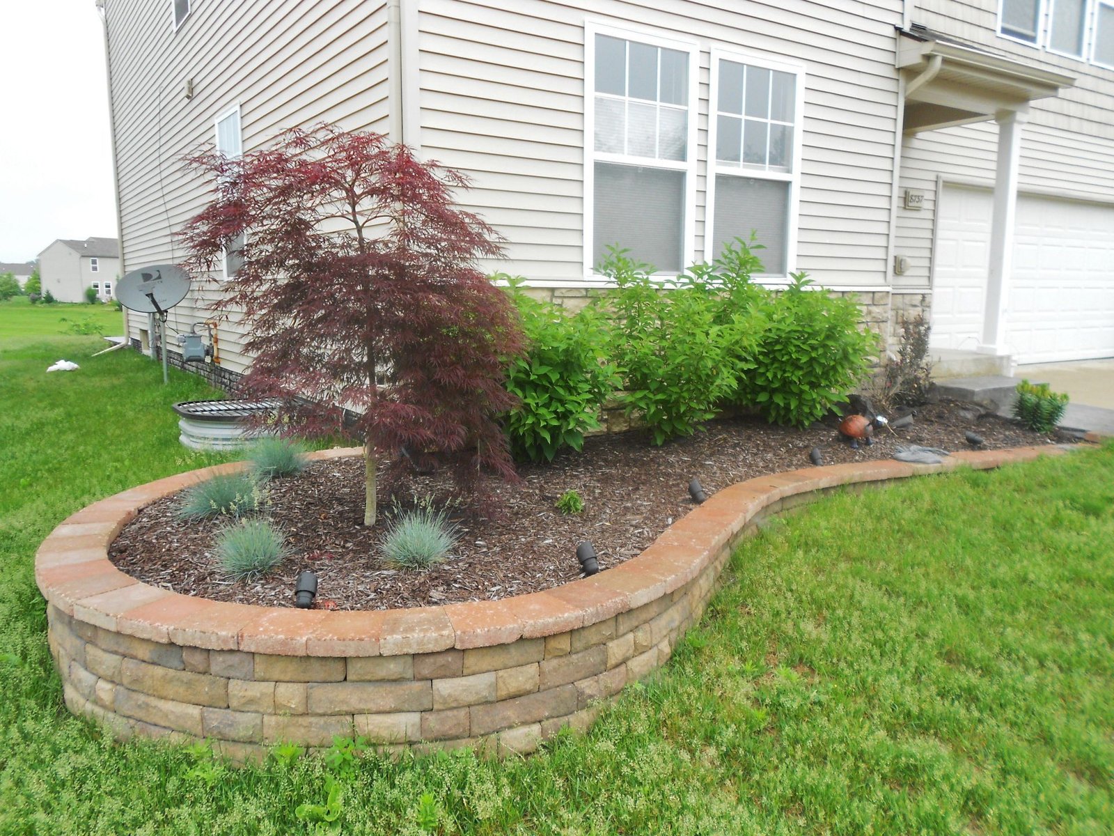 Front yard landscaping with a curved stone flower bed, red and green shrubs beside a house and garage.