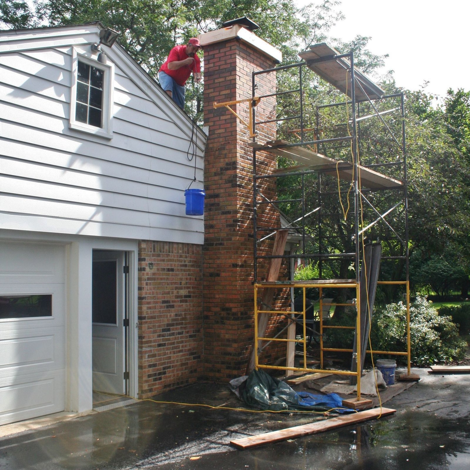 Worker on scaffolding repairing a brick chimney beside a house with an open garage.