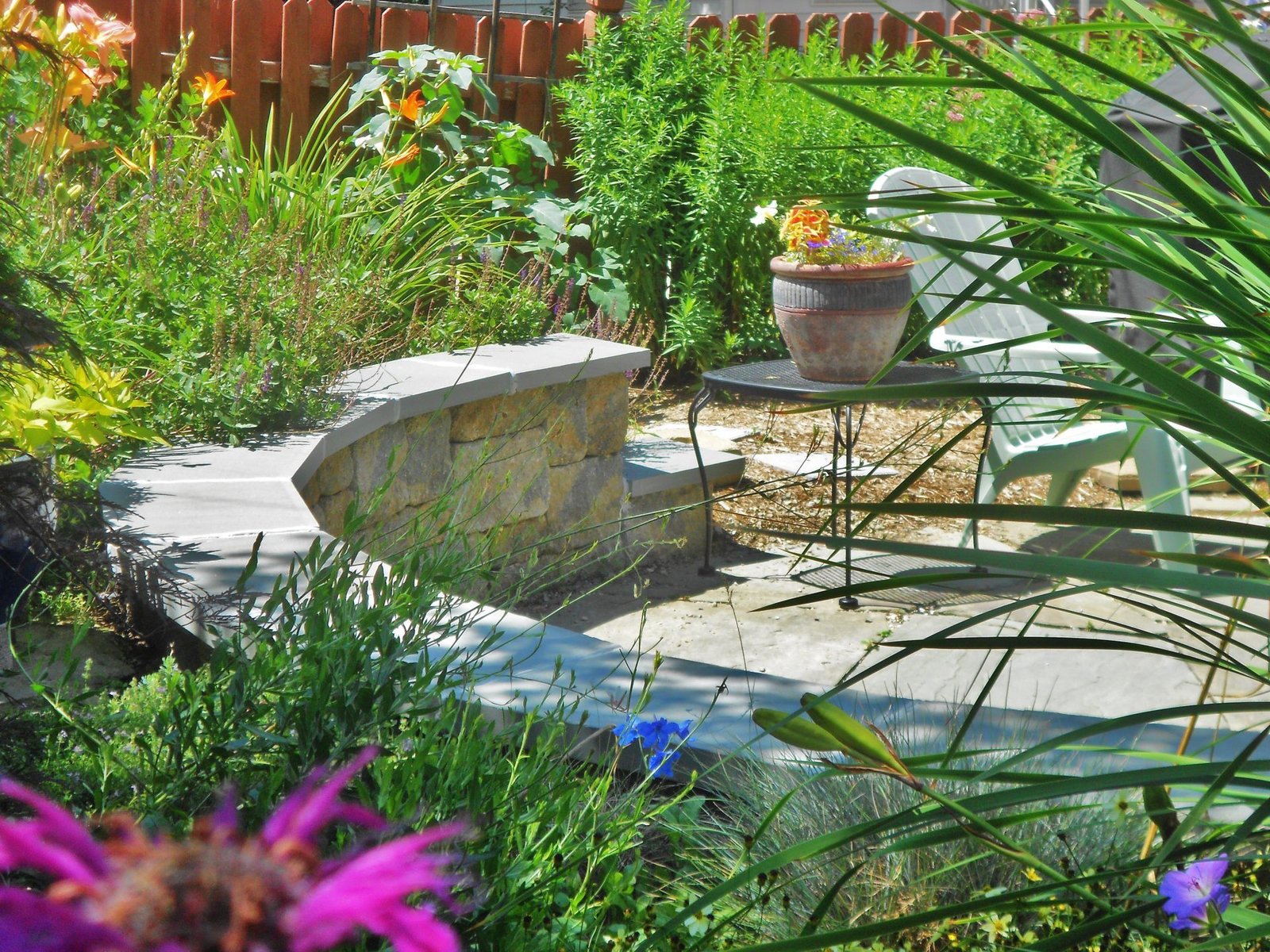Sunny garden patio with stone bench, metal table, potted flowers, and lush green plants