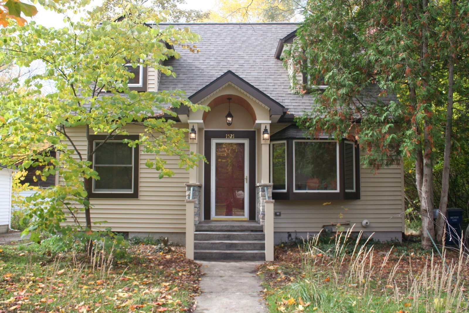 Small beige house with black shutters, a front porch, and a walkway surrounded by trees and garden plants