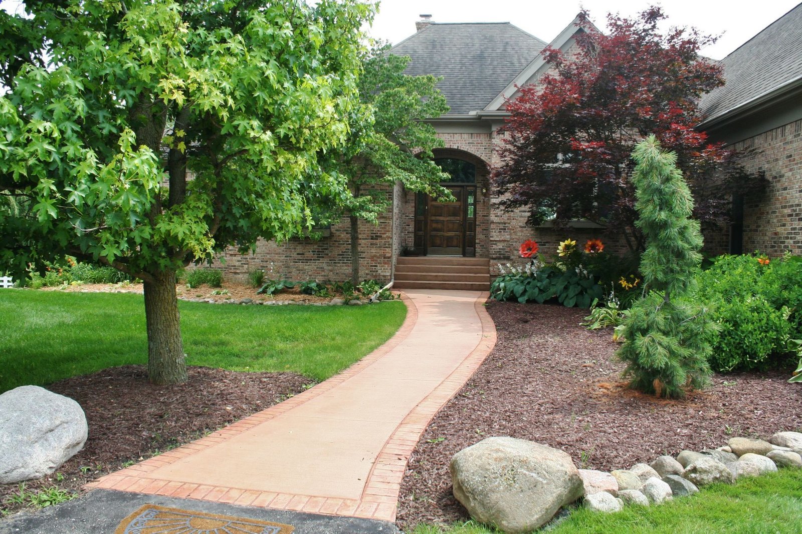 Curved brick walkway leading to a house entrance, bordered by green trees, shrubs, and rocks