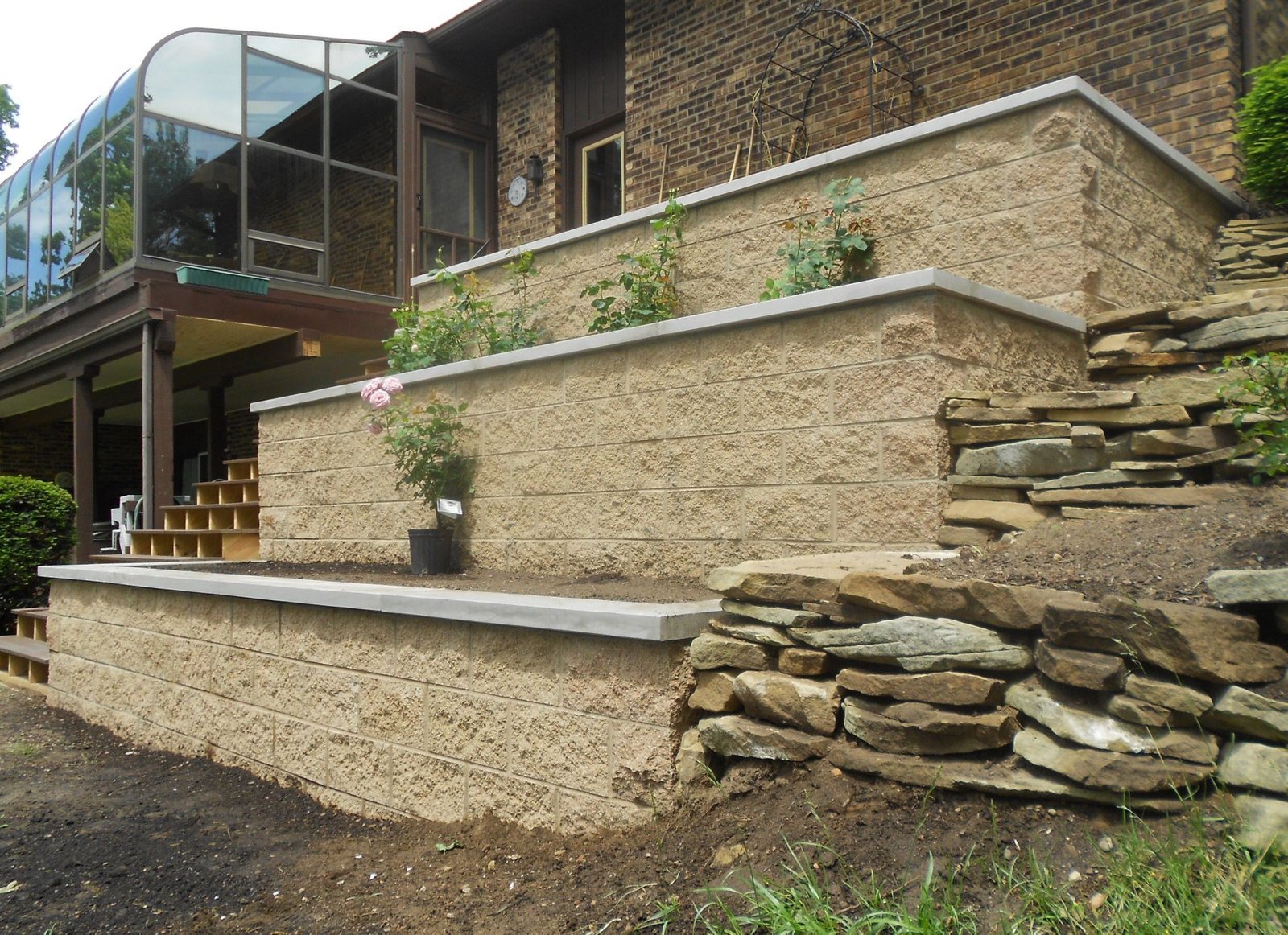 Tiered concrete block retaining walls with capstones and a dry-stack stone corner beside a brick home